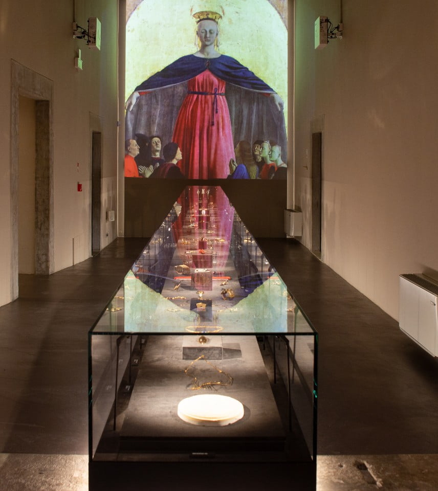 Immagine di una delle sale del Museo Orodautore, con gioielli in oro racchiusi in un lungo espositore orizzontale. Sullo sfondo proiezione multimediale della "Madonna della Misericordia" di Piero della Francesca.
