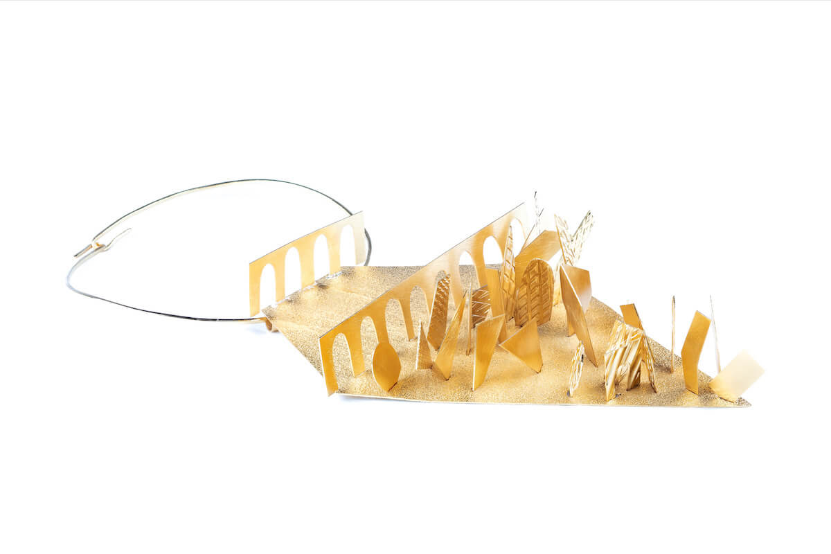 Collare scultoreo con anello metallico e piastra dorata “pop-up” che forma archi e elementi geometrici, come un paesaggio astratto.