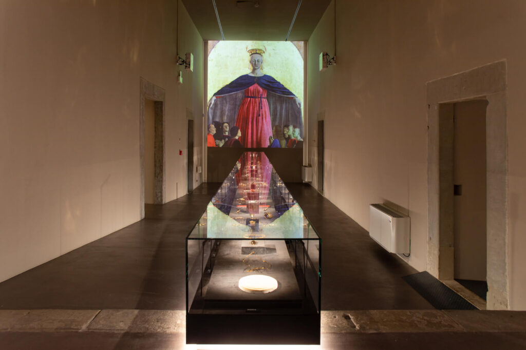 Immagine di una delle sale del Museo Orodautore, con gioielli in oro racchiusi in un lungo espositore orizzontale. Sullo sfondo proiezione multimediale della "Madonna della Misericordia" di Piero della Francesca.